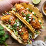 Crispy hot honey garlic chicken tacos held in hand with fresh cabbage slaw, cotija cheese, and cilantro